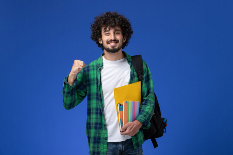 a1front view male student wearing black backpack holding copybooks files blue wall